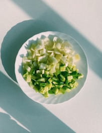 Chopped green vegetables, possibly celery and bell peppers, are arranged on a white plate placed on a light-colored surface. The scene is lit by natural sunlight, creating distinct shadows that stretch across the surface.