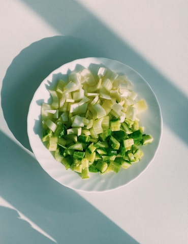 Chopped green vegetables, possibly celery and bell peppers, are arranged on a white plate placed on a light-colored surface. The scene is lit by natural sunlight, creating distinct shadows that stretch across the surface.