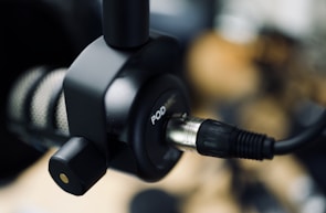 A close-up of a professional podcast microphone with a cable plugged into it. The focus is on the connector and part of the microphone, with a blurred background.