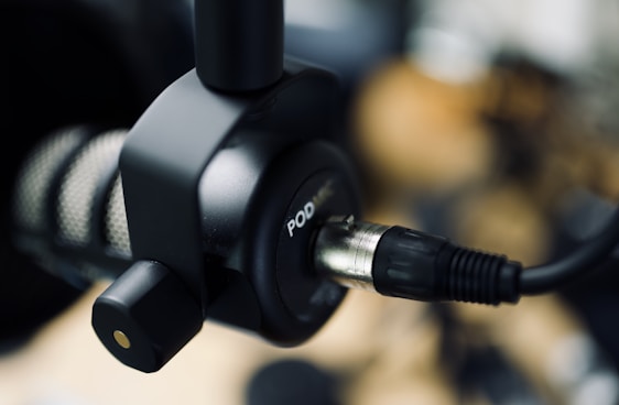 A close-up of a professional podcast microphone with a cable plugged into it. The focus is on the connector and part of the microphone, with a blurred background.
