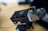 A close-up of a portable audio recorder, featuring a display screen and several buttons including a stop and play button. The device rests slightly tilted, possibly mounted on a stand, with visible knobs and microphone inputs.