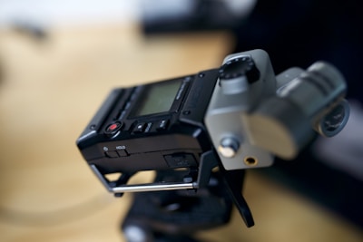 A close-up of a portable audio recorder, featuring a display screen and several buttons including a stop and play button. The device rests slightly tilted, possibly mounted on a stand, with visible knobs and microphone inputs.