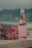 A glass bottle of Hatsu soda with a pink liquid is placed on a wooden surface, accompanied by an arrangement of vibrant flowers in shades of pink, purple, and yellow. The background features a blurred landscape with greenery and overcast skies.