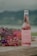 A glass bottle of Hatsu soda with a pink liquid is placed on a wooden surface, accompanied by an arrangement of vibrant flowers in shades of pink, purple, and yellow. The background features a blurred landscape with greenery and overcast skies.
