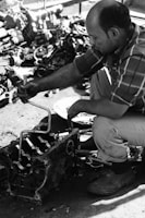 A person is working on a mechanical device, possibly an engine, using a hand tool. The person is wearing a plaid shirt and pants, and the environment appears to be a workshop with various mechanical parts scattered on the ground.