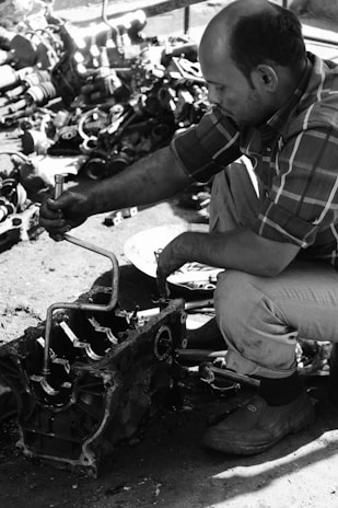 A person is working on a mechanical device, possibly an engine, using a hand tool. The person is wearing a plaid shirt and pants, and the environment appears to be a workshop with various mechanical parts scattered on the ground.