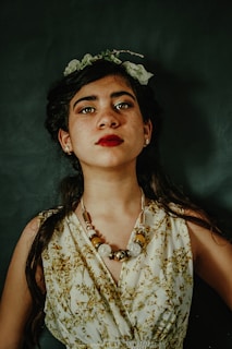 A young woman with dark hair gazes confidently at the camera. She is adorned with a floral headpiece and a bold beaded necklace. Her attire features a pattern of green and brown botanical prints. The background is dark, enhancing her striking red lipstick and the details of her outfit.