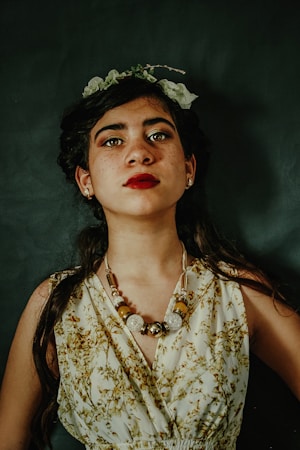 A young woman with dark hair gazes confidently at the camera. She is adorned with a floral headpiece and a bold beaded necklace. Her attire features a pattern of green and brown botanical prints. The background is dark, enhancing her striking red lipstick and the details of her outfit.