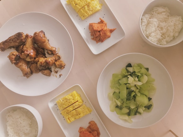 A variety of dishes arranged on a table, including fried chicken pieces, scrambled eggs, kimchi, white rice, and a bowl of steamed green vegetables.