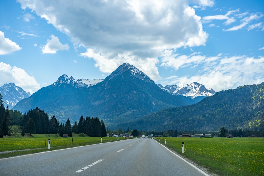 camino de asfalto gris cerca del campo de hierba verde y árboles verdes y montañas bajo nubes blancas y