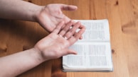 A pair of open hands held above an open book, which appears to be a Bible, on a wooden table.