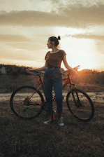 woman in brown tank top and blue denim jeans standing beside black bicycle during sunset