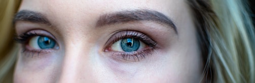 A close-up of a person's blue eyes with light makeup, emphasizing the lashes and brows. The skin tone is fair, and hair is visible at the edges.