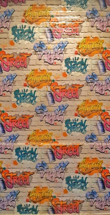 A vibrant graffiti mural covers a brick wall. Various words like 'Spray', 'Street', and 'Swag' are depicted in bright colors, including yellow, pink, blue, orange, and green, with artistic flair and dynamic styles. The background features a realistic brick texture with splashes of color surrounding the text.