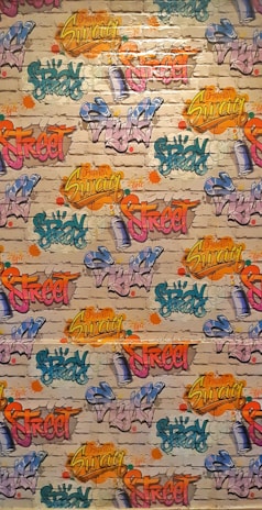 A vibrant graffiti mural covers a brick wall. Various words like 'Spray', 'Street', and 'Swag' are depicted in bright colors, including yellow, pink, blue, orange, and green, with artistic flair and dynamic styles. The background features a realistic brick texture with splashes of color surrounding the text.