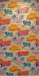 A vibrant graffiti mural covers a brick wall. Various words like 'Spray', 'Street', and 'Swag' are depicted in bright colors, including yellow, pink, blue, orange, and green, with artistic flair and dynamic styles. The background features a realistic brick texture with splashes of color surrounding the text.