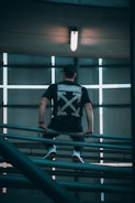 A person is perched on a metal railing in an industrial setting with a large light overhead. The back of their black t-shirt features a bold graphic design with arrows. The image has a symmetrical composition with linear elements and subdued lighting.