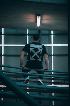 A person is perched on a metal railing in an industrial setting with a large light overhead. The back of their black t-shirt features a bold graphic design with arrows. The image has a symmetrical composition with linear elements and subdued lighting.