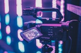 A professional video camera set on a tripod is surrounded by a vibrant display of pink and blue neon lights. The camera's viewfinder shows a digital display, indicating that it is turned on and possibly recording. The background features blurred vertical patterns of colored lights, creating a modern and dynamic atmosphere.