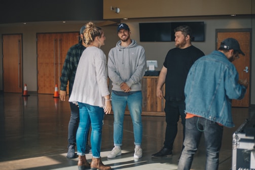 A group of five people are casually gathered in a spacious, well-lit room with wooden doors and flat screen monitors mounted on the wall. The individuals are dressed in casual attire, such as jeans, hoodies, and jackets. They appear to be engaged in a relaxed, friendly conversation.