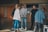 A group of five people are casually gathered in a spacious, well-lit room with wooden doors and flat screen monitors mounted on the wall. The individuals are dressed in casual attire, such as jeans, hoodies, and jackets. They appear to be engaged in a relaxed, friendly conversation.