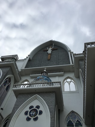 A large church building is depicted, showcasing intricate Gothic architecture. A prominent crucifix with a statue of Jesus is mounted above the entrance. The facade includes ornate stained glass windows and decorative arches. The sky is overcast, adding a subdued lighting to the scene.