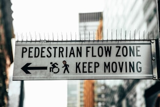 A metal sign prominently displays the words 'PEDESTRIAN FLOW ZONE' and 'KEEP MOVING' in black capital letters. The sign includes a left-pointing arrow and icons indicating directions for movement, with a protective row of spikes on top. The background features blurred urban architecture.