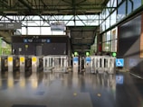 A modern transit station entrance with several automated turnstiles, featuring a combination of metal and glass materials. Signs above the turnstiles indicate directional arrows and possible station information. The station has a wheelchair accessible gate, marked by a blue sign with a wheelchair symbol. Sunlight streams through large glass windows, casting shadows on the polished floor.