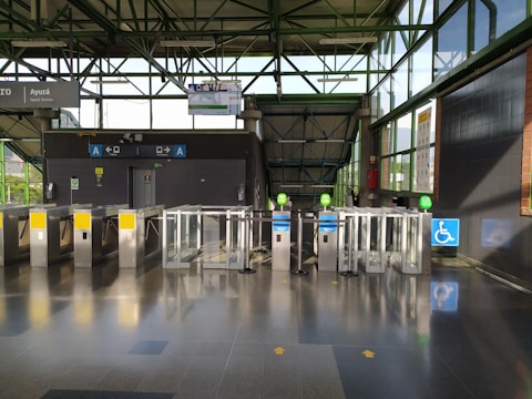 A modern transit station entrance with several automated turnstiles, featuring a combination of metal and glass materials. Signs above the turnstiles indicate directional arrows and possible station information. The station has a wheelchair accessible gate, marked by a blue sign with a wheelchair symbol. Sunlight streams through large glass windows, casting shadows on the polished floor.