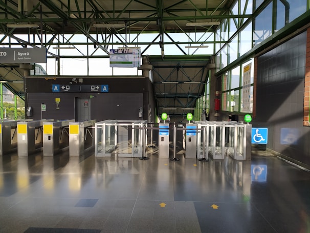 A modern transit station entrance with several automated turnstiles, featuring a combination of metal and glass materials. Signs above the turnstiles indicate directional arrows and possible station information. The station has a wheelchair accessible gate, marked by a blue sign with a wheelchair symbol. Sunlight streams through large glass windows, casting shadows on the polished floor.