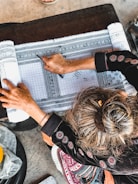 A person with intricately designed traditional clothing is engaged in a detailed drawing or writing activity on patterned fabric. The hand is holding a tool, and the focus is on the precision of the pattern work. The surface is wooden, and the environment suggests a workspace dedicated to craft or art.