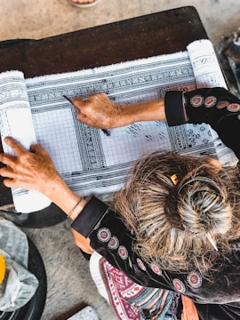 A person with intricately designed traditional clothing is engaged in a detailed drawing or writing activity on patterned fabric. The hand is holding a tool, and the focus is on the precision of the pattern work. The surface is wooden, and the environment suggests a workspace dedicated to craft or art.