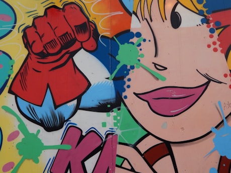 A vibrant graffiti artwork featuring a comic-style depiction of a close-up female face with bold lipstick and an exaggerated eye. A red-gloved fist is prominent, suggesting a superhero motif. The background is filled with colorful splashes and dots in shades of blue, green, pink, and orange.