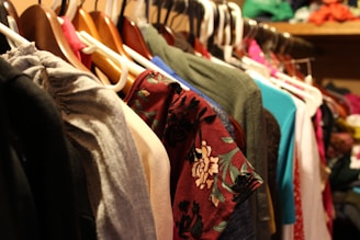 A variety of clothes hang on a set of hangers in a closet. The garments are of different colors and patterns, with some featuring floral designs. The hangers are primarily plastic and wood, and the surrounding area suggests a crowded and organized storage space.