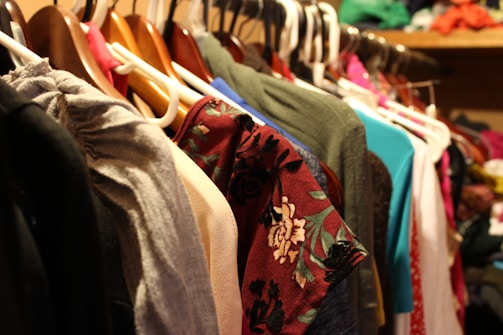 A variety of clothes hang on a set of hangers in a closet. The garments are of different colors and patterns, with some featuring floral designs. The hangers are primarily plastic and wood, and the surrounding area suggests a crowded and organized storage space.