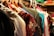 A variety of clothes hang on a set of hangers in a closet. The garments are of different colors and patterns, with some featuring floral designs. The hangers are primarily plastic and wood, and the surrounding area suggests a crowded and organized storage space.
