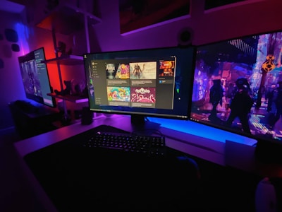 A gaming setup featuring multiple screens with vibrant game-related images illuminated by colorful LED lighting. The environment is dimly lit, highlighting the vivid displays on the monitors. A keyboard is visible on the desk beneath the central monitor.