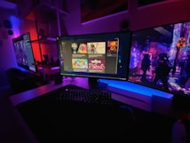 A gaming setup featuring multiple screens with vibrant game-related images illuminated by colorful LED lighting. The environment is dimly lit, highlighting the vivid displays on the monitors. A keyboard is visible on the desk beneath the central monitor.
