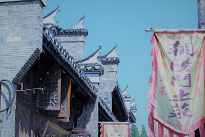 A traditional Chinese architectural setting with stone buildings featuring ornate roof designs. Brightly colored banners with Chinese characters hang prominently, adding vibrance to the scene.
