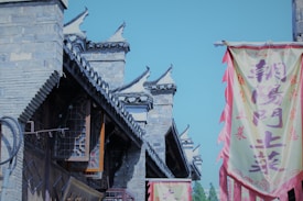 A traditional Chinese architectural setting with stone buildings featuring ornate roof designs. Brightly colored banners with Chinese characters hang prominently, adding vibrance to the scene.