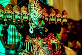 A person is dressed in an elaborate costume with multiple decorated headdresses, each featuring a painted face. The outfit is richly adorned with embroidery and jewels, showcasing colorful and intricate patterns. The background contains blurred figures, creating a dynamic and festive atmosphere.