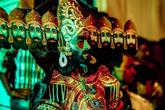 A person is dressed in an elaborate costume with multiple decorated headdresses, each featuring a painted face. The outfit is richly adorned with embroidery and jewels, showcasing colorful and intricate patterns. The background contains blurred figures, creating a dynamic and festive atmosphere.