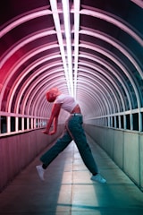 A person is performing an expressive, dynamic dance in the middle of an illuminated tunnel with arched metal framework. The individual is mid-air, with arms extended backward and the body in a curved, graceful pose, wearing a white crop top, green pants, and white sneakers. The tunnel is lit with vibrant red and blue neon lights that create a futuristic ambiance.