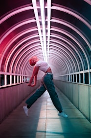 A person is performing an expressive, dynamic dance in the middle of an illuminated tunnel with arched metal framework. The individual is mid-air, with arms extended backward and the body in a curved, graceful pose, wearing a white crop top, green pants, and white sneakers. The tunnel is lit with vibrant red and blue neon lights that create a futuristic ambiance.