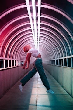 A person is performing an expressive, dynamic dance in the middle of an illuminated tunnel with arched metal framework. The individual is mid-air, with arms extended backward and the body in a curved, graceful pose, wearing a white crop top, green pants, and white sneakers. The tunnel is lit with vibrant red and blue neon lights that create a futuristic ambiance.