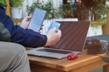 A person is using a smartphone and holding a credit card in front of a laptop on a wooden table. To the side, there is a digital camera and a red electronic device, possibly a pair of wireless earbuds in a case. Indoor plants are visible in the background, adding a touch of greenery.