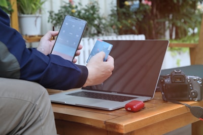 A person is using a smartphone and holding a credit card in front of a laptop on a wooden table. To the side, there is a digital camera and a red electronic device, possibly a pair of wireless earbuds in a case. Indoor plants are visible in the background, adding a touch of greenery.
