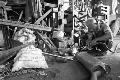 A man is engaged in welding in a cluttered workshop. Surrounding him are various metal parts, tools, and bags. Sparks are visibly flying from the welding process. The environment appears industrial and is filled with disorganized equipment.