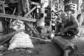 A man is engaged in welding in a cluttered workshop. Surrounding him are various metal parts, tools, and bags. Sparks are visibly flying from the welding process. The environment appears industrial and is filled with disorganized equipment.
