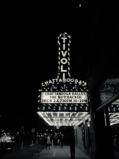 A black and white photograph of a theater entrance with a marquee that reads 'CHATTANOOGA BALLET THE NUTCRACKER DEC 9 2 & 7:30PM 10-2PM' in illuminated letters. The large signage of the Tivoli Theatre is vertical and well-lit. People walk underneath the marquee on the sidewalk, and the street is lined with lights and signs.